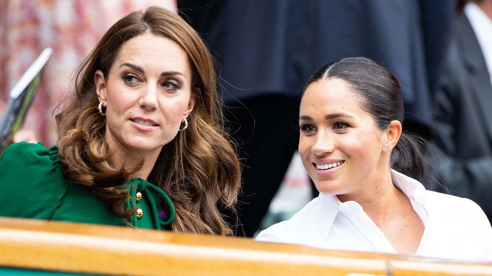 Kate Middleton and Meghan Markle sit and talk together looking to their right, as Markle smiles