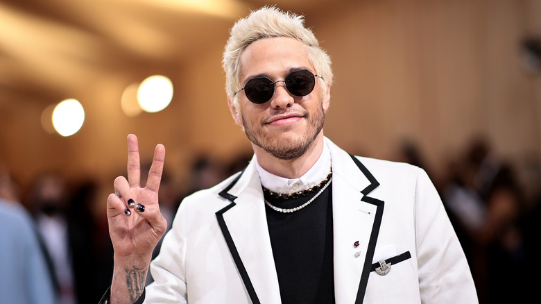 Pete Davidson wearing a white suit and round black sunglasses, holding his hand in a peace sign