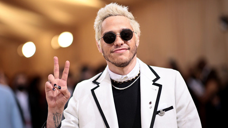 Pete Davidson wearing a white suit and round black sunglasses, holding his hand in a peace sign