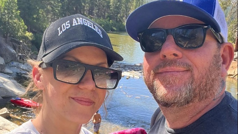 Jodie Sweetin and Mescal Wasilewski wearing hats and sunglasses and taking a selfie beside a river.