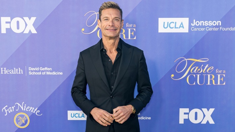 Ryan Seacrest wears a black suit jacket and matching shirt while posing with his hands in front of him.
