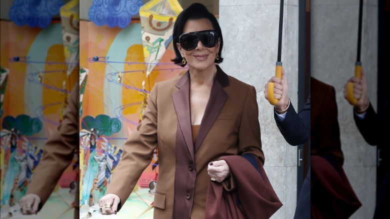 Kris Jenner walking outdoors and wearing sunglasses as someone holds an umbrella over her