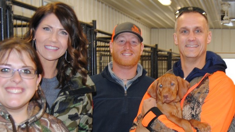Corey Lewandowski holding a puppy at a kennel while posing with Kristi Noem and two other people