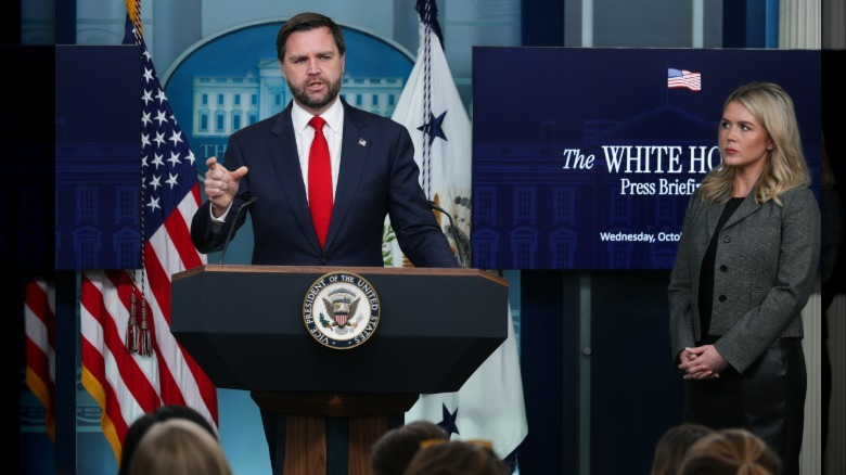 Karoline Leavitt standing to the side and watching JD Vance address the press from the lectern in the White House briefing room